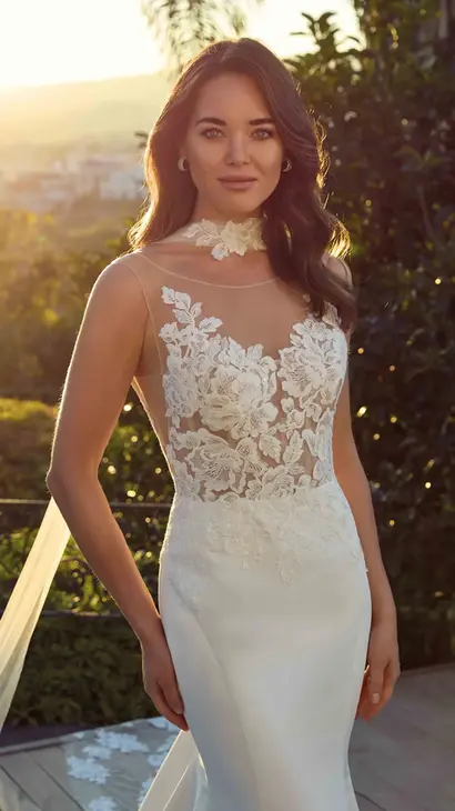 Close up of Eddy K satin mermaid wedding dress with an illusion neckline and a sleeveless body on a bridal model standing in a garden with a sunset as a background.