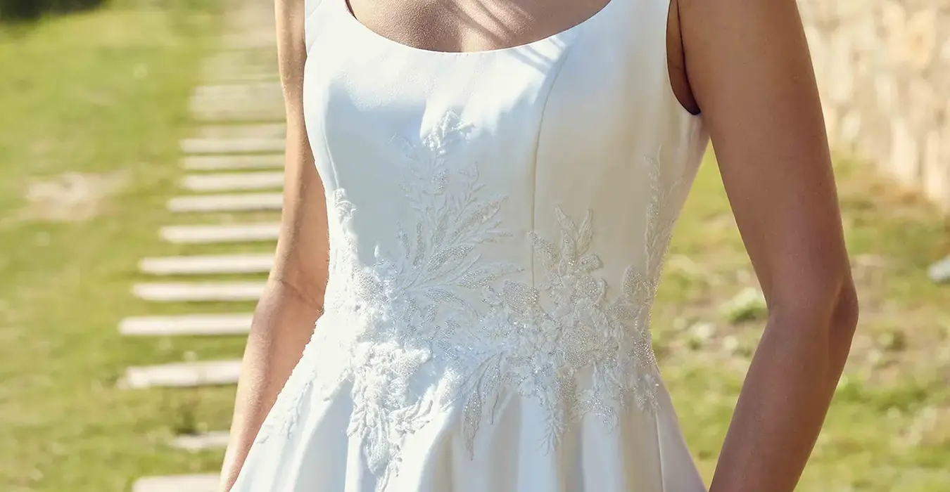 Close up of Eddy K satin ballgown wedding dress with a scoop neckline and a sleeveless body on a bridal model posing on a park in a sunny day with green grass and threes in the background.
