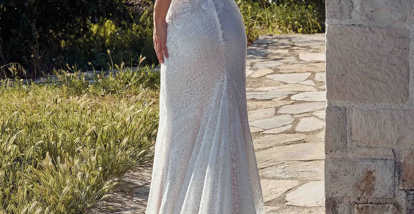 Back of Eddy K all over lace fit-n-flare wedding dress with key-hole back and open train on bride standing in rustic garden.
