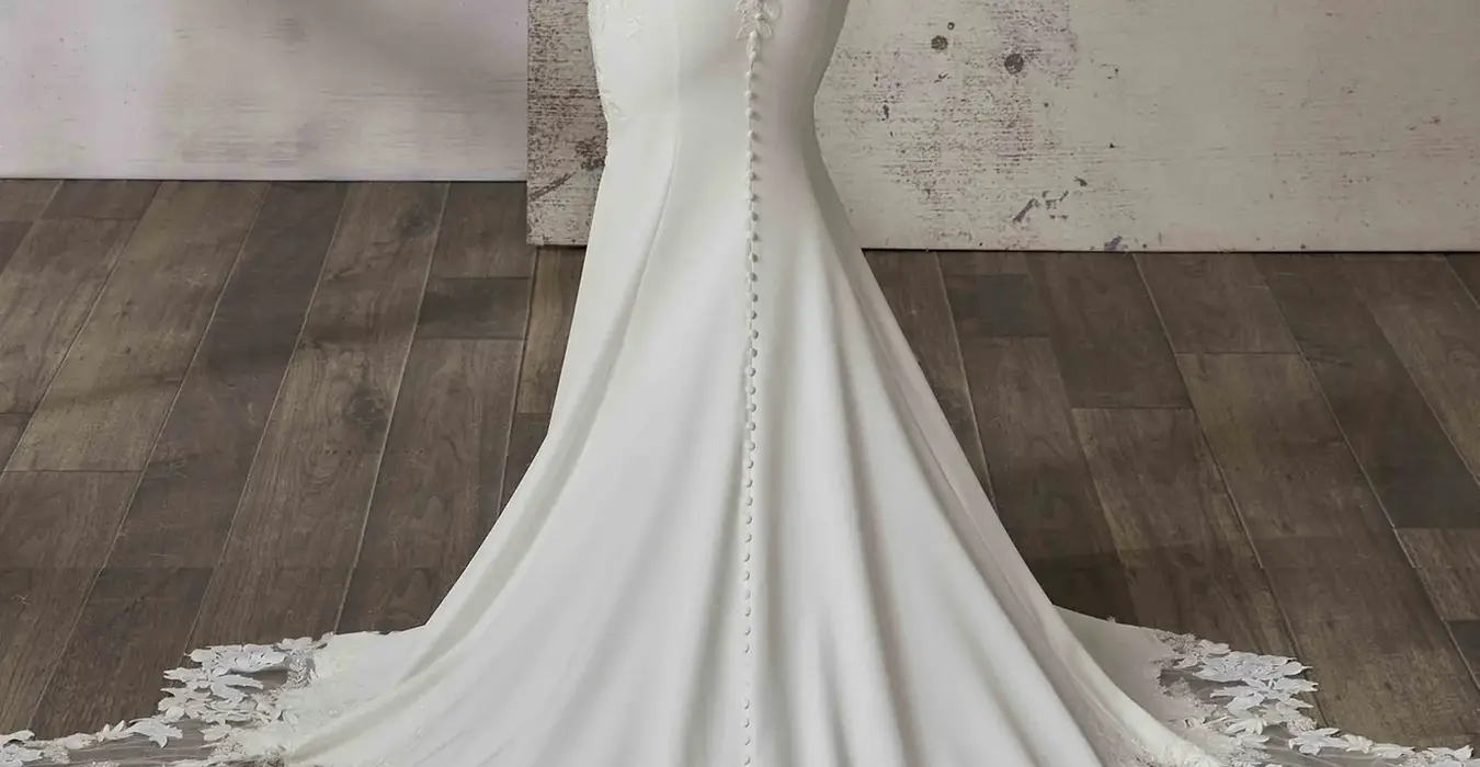 Back of Eddy K crepe fit-n-flare wedding dress with skin unlined back and a big illusion train on bride standing in studio.