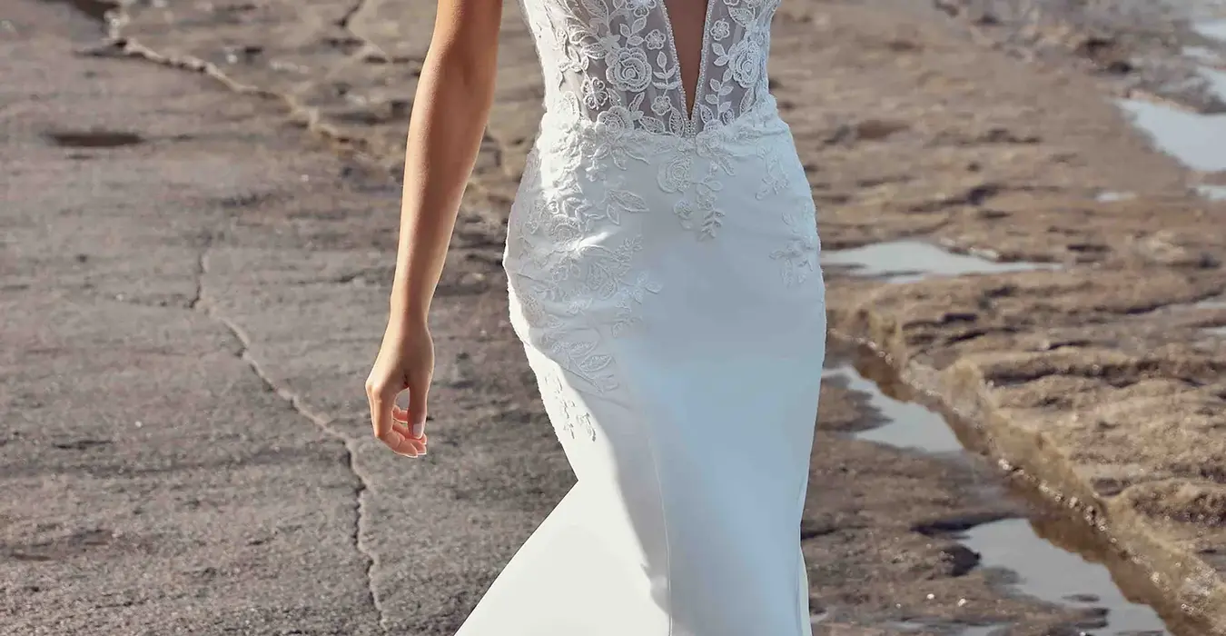 Eddy K fit-n-flare crepe and lace with spaghetti straps and deep v neckline on a bridal model posing with hand in her hair on the beach.