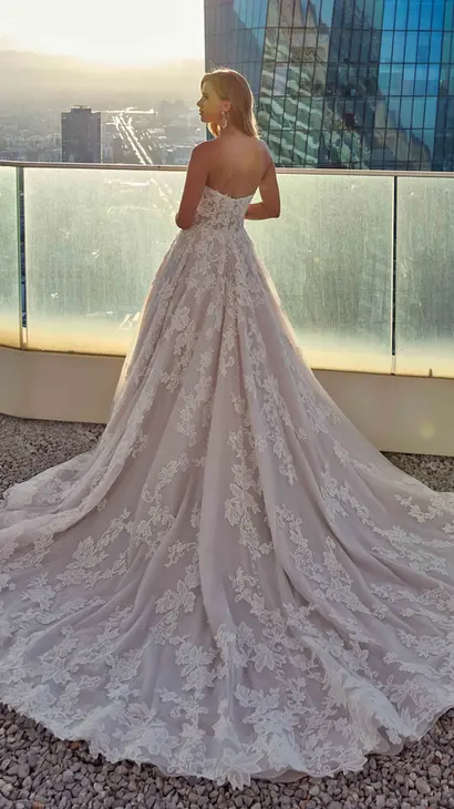 Back Eddy K lace a-line wedding dress with a straight neckline and a strapless body on a bridal model standing on a rooftop of a skyscraper with a nice sunset and a distant view of the city and other buildings in the background
