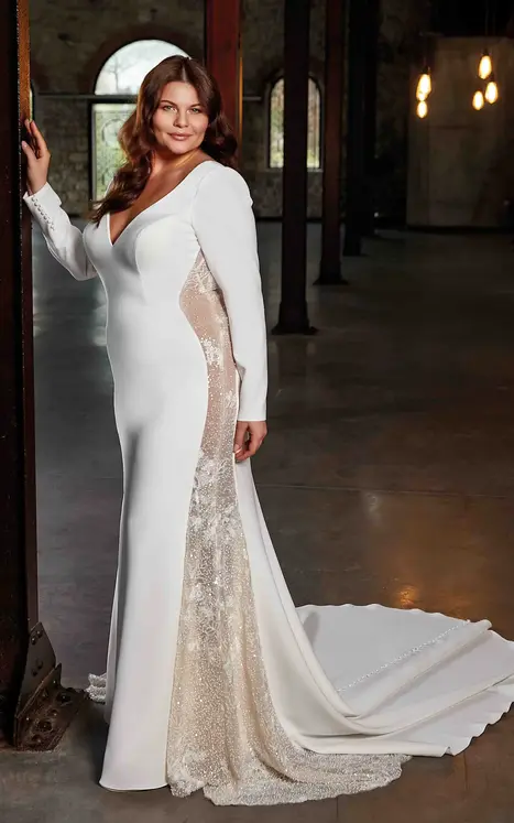 Side view of Eddy K crepe long sleeve wedding dress with side panels of pearl beaded embroidery and sequins over tulle o bridal model standing next to wood beam.