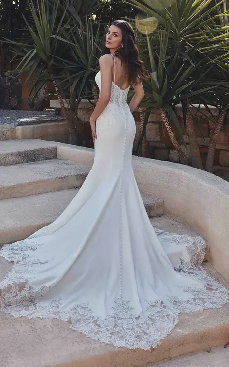 Back of Eddy K crepe fit-n-flare wedding dress with lace cut-out train and spaghetti straps on bridal model standing on stairs with palm trees in background.