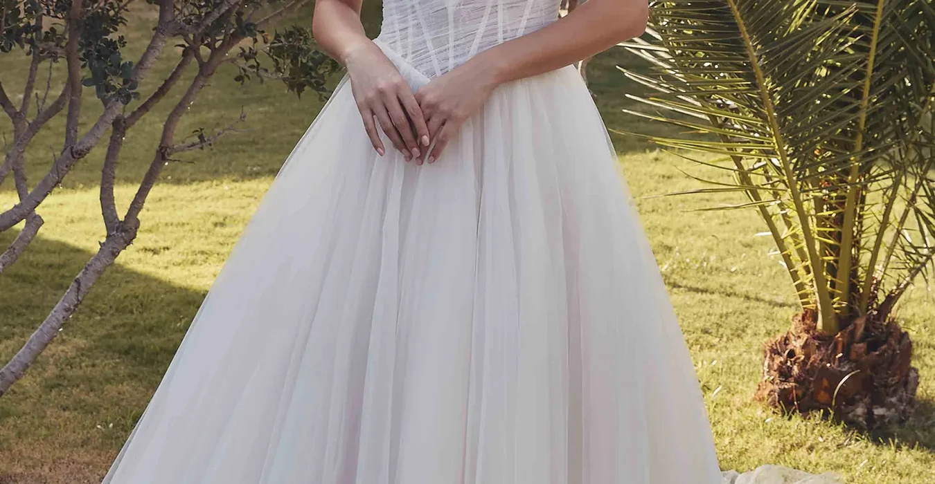 Eddy K tulle ballgown wedding dress with basque waist and off shoulder swag straps on bridal model standing in greenery garden.