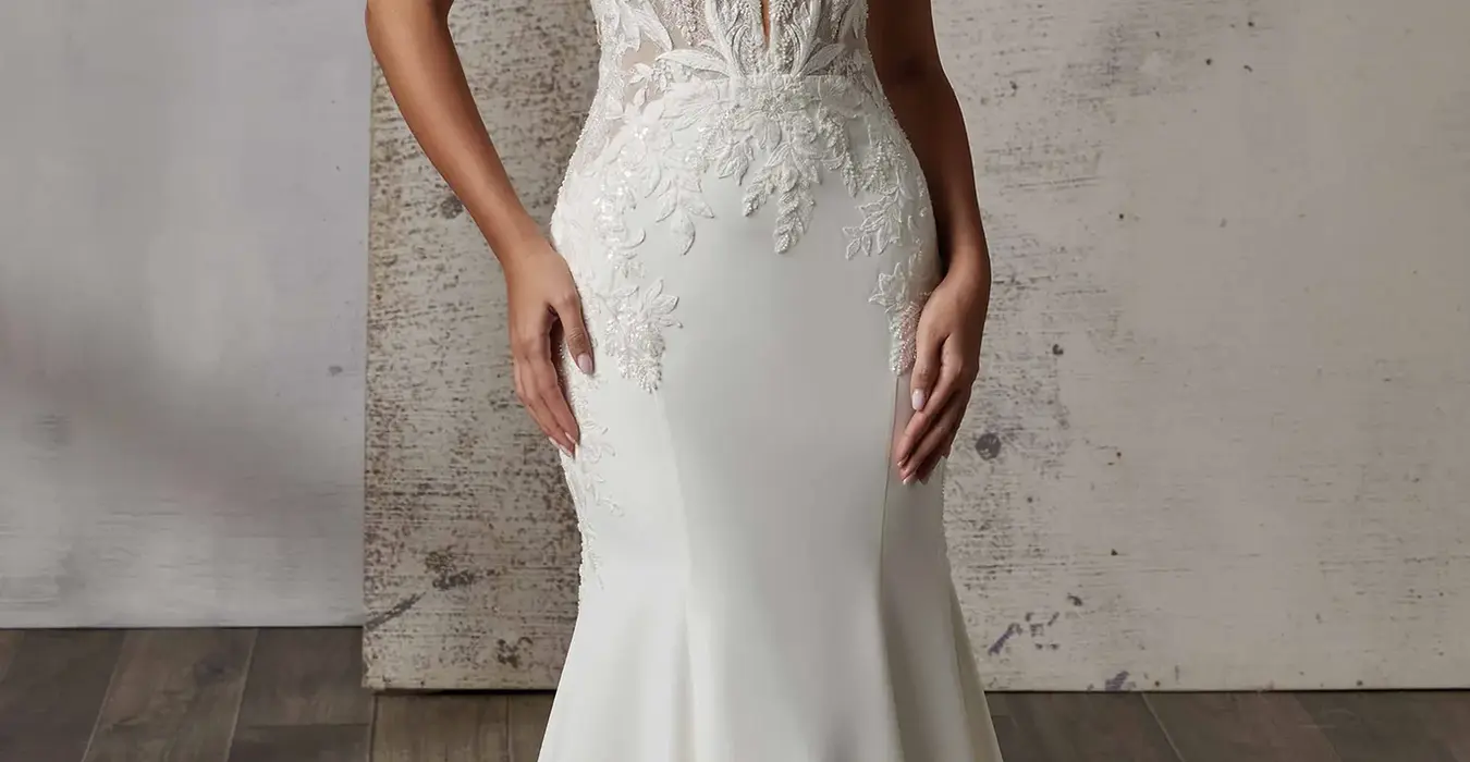 Eddy K fit-n-flare crepe wedding gown with spaghetti and lace unlined bodice and cut out lace train on bridal model standing in studio