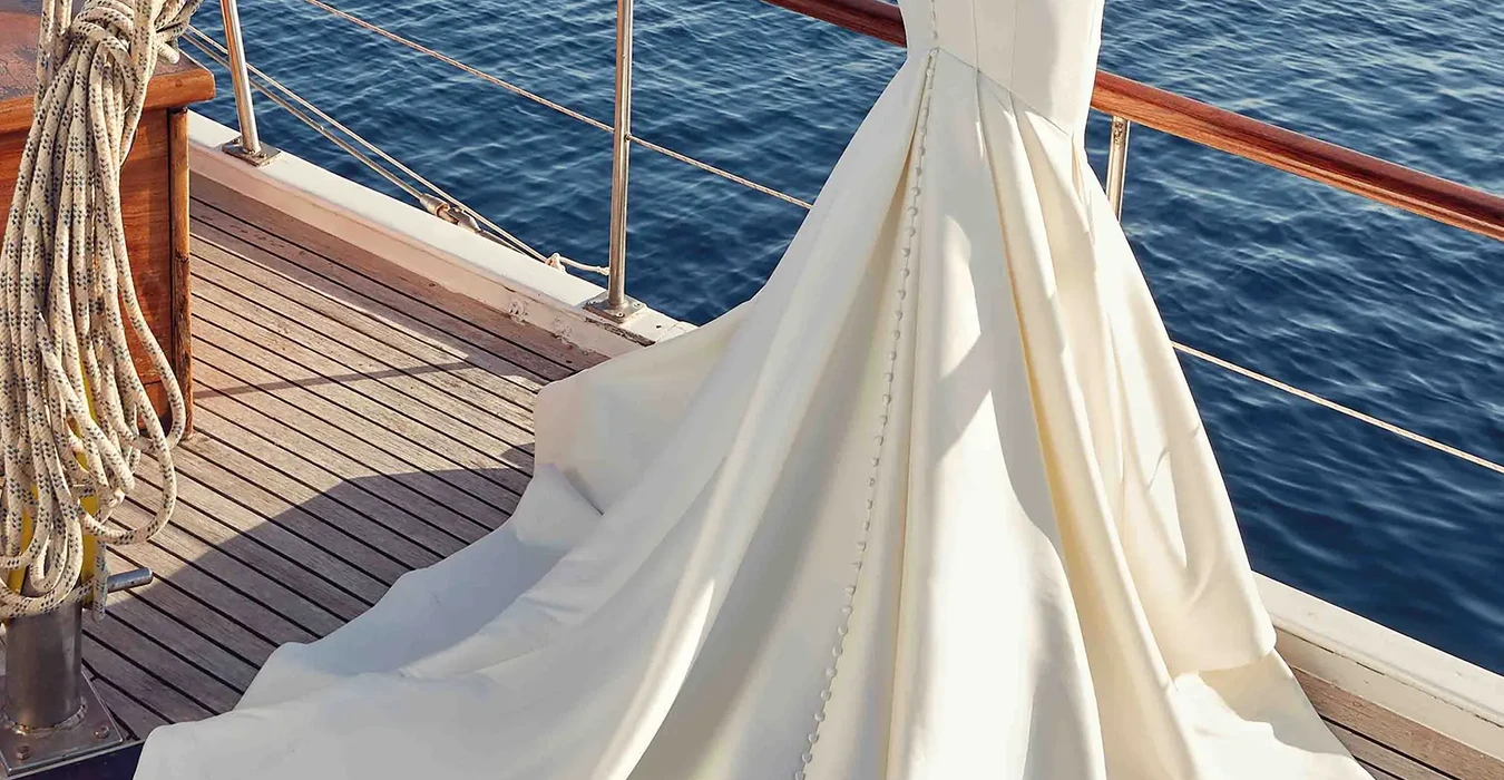 Back of Eddy K strapless mikado mermaid wedding gown with illusion cut-outs in the back on bride standing on a boat in the ocean.