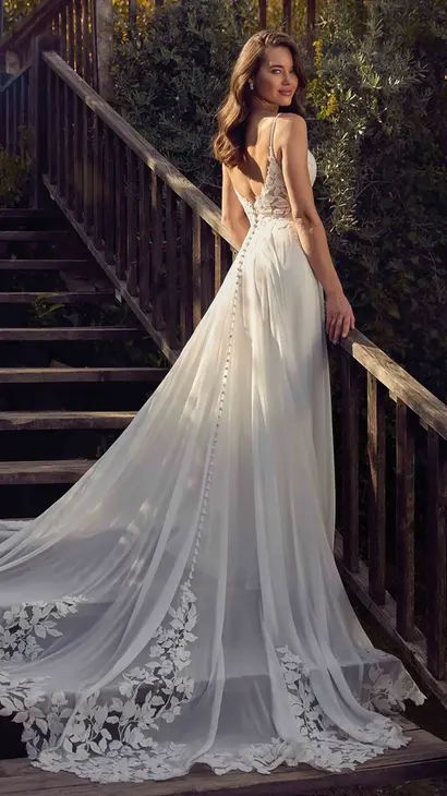 Back of Eddy K chiffon a-line wedding dress with a v-neck neckline and a sleeveless body on a bridal model posing in front of wooden stairs from a terrace in a garden.