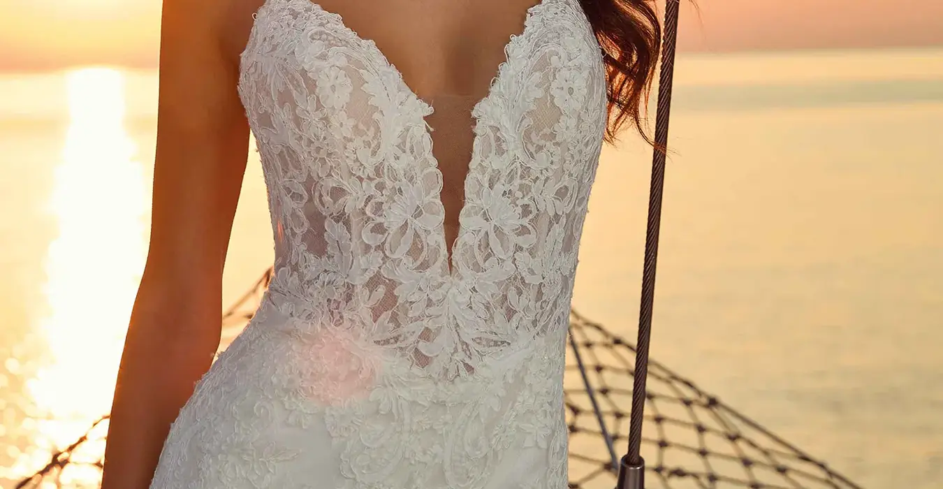 Close up of Eddy K lace fit-n-flare wedding dress with a v-neck neckline and a sleeveless body on a bridal model posing on a yacht with a beautiful sunset in the background.