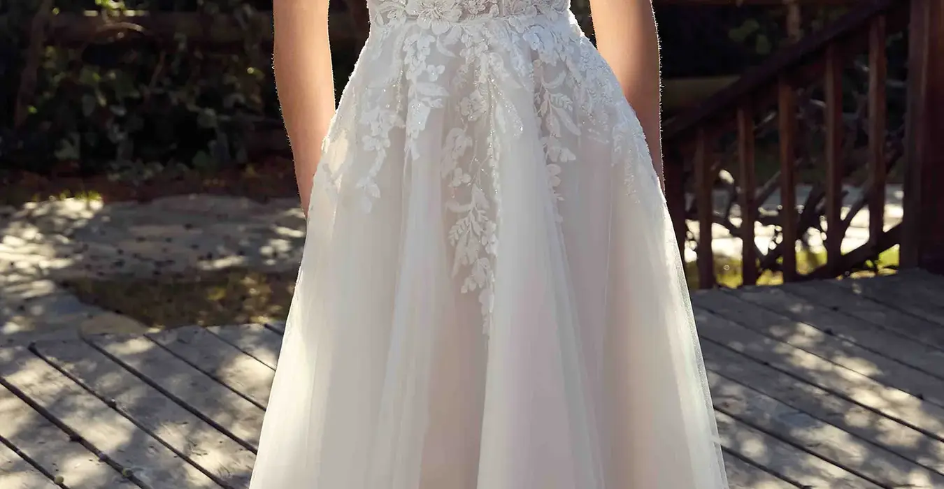 Eddy K lace mermaid wedding dress with a v-neck neckline and a sleeveless body on a bridal model posing in a park in a sunny day with threes in the background.