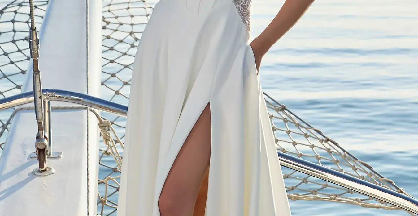 Eddy K crêpe a-line wedding dress with a scoop neckline and a sleeveless body on a bridal model posing on a yacht on a sunny day where the sea can be seen in the background.