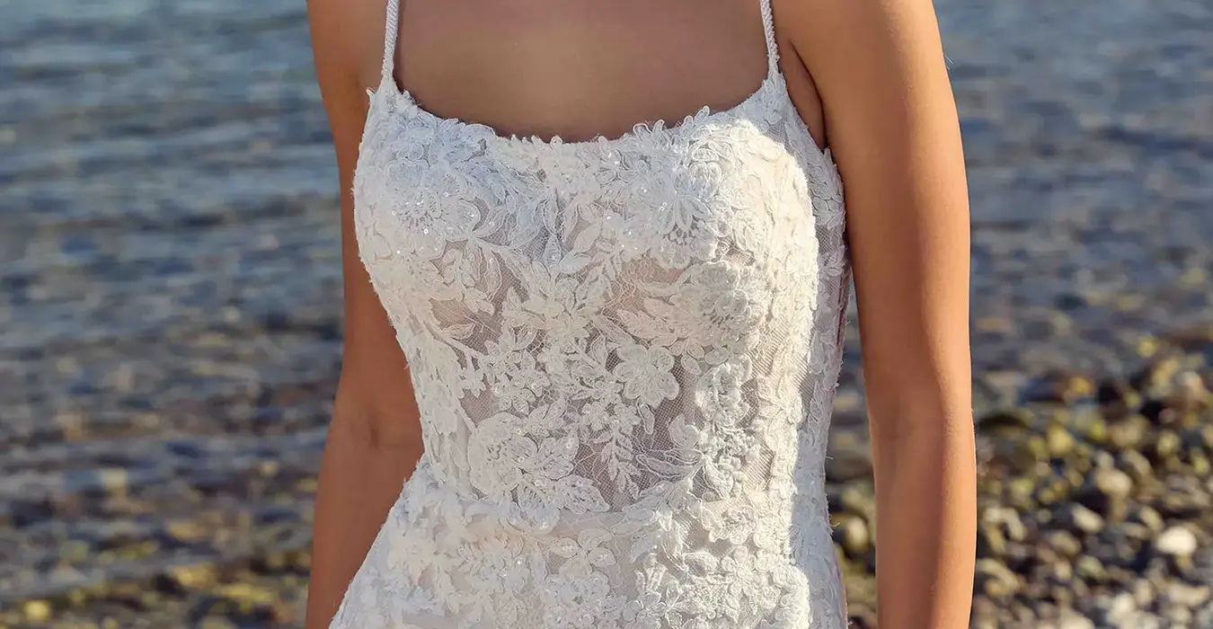 Close up of Eddy K lace fit-n-flare wedding dress with a straight neckline and a sleeveless body on a bridal model standing on a rocky beach.