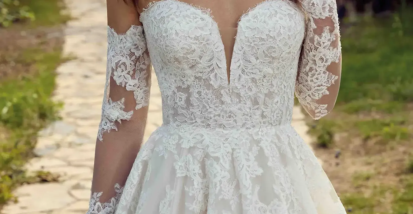 Close up of Eddy K tulle/lace ballgown wedding dress with a sweetheart neckline and a long sleeves body on a bridal model posing in a nice garden surrounded by threes.