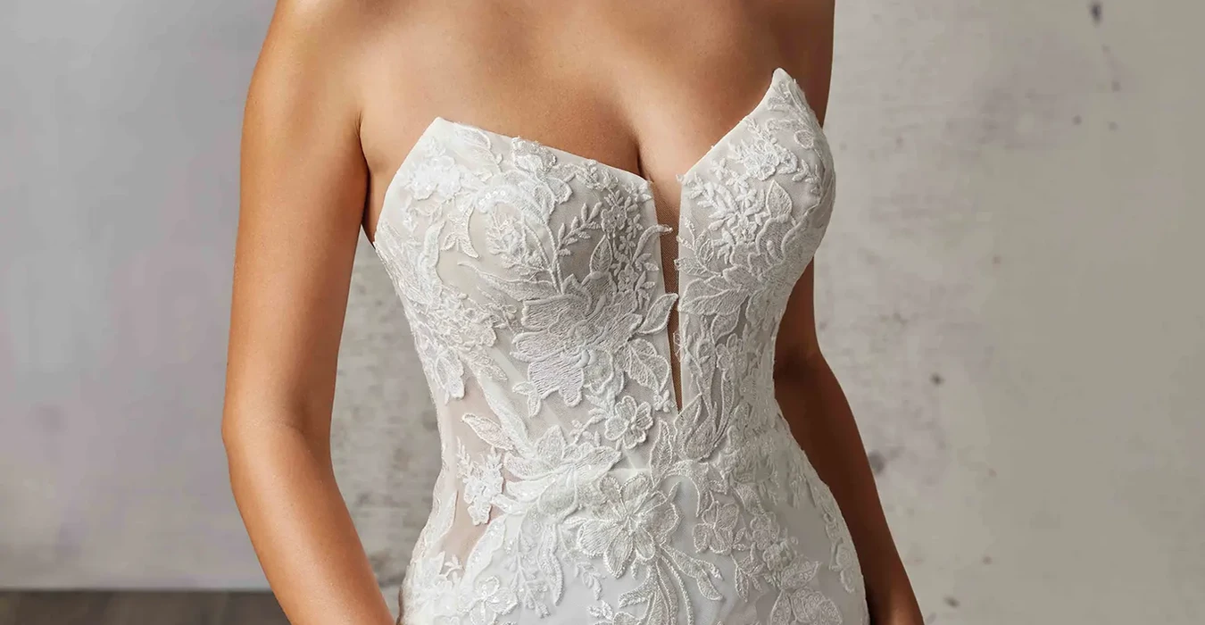 Close up of Eddy K strapless all over lace wedding dress with cat-eye neckline and skin cleavage on bridal model standing in studio.