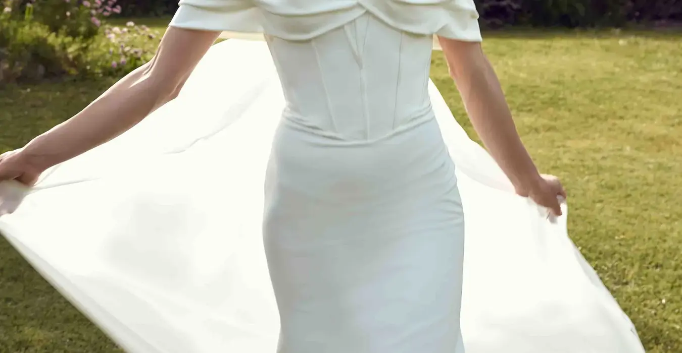 Eddy K crepe fit-n-flare wedding dress with a sweetheart neckline and an off-shoulder bodice on a bridal model standing in a garden