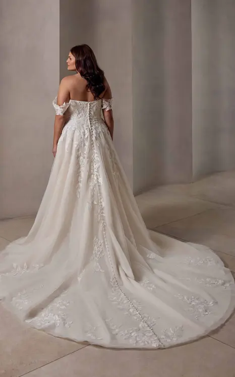 Back of Eddy K lace d tulle ballgown wedding dress with a sweetheart neckline and off-shoulder swag slleves on a bridal model standing in an elegant studio.