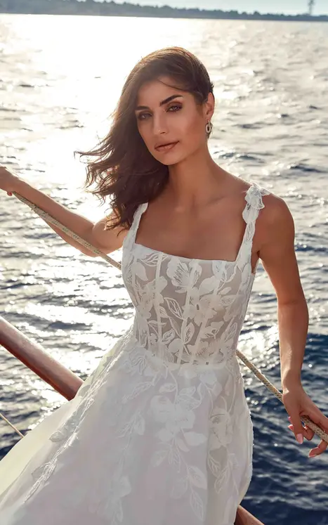 Close up of Eddy K organza and lace wedding gown with square neckline and thick lace straps worn by bride sitting on edge of a boat.