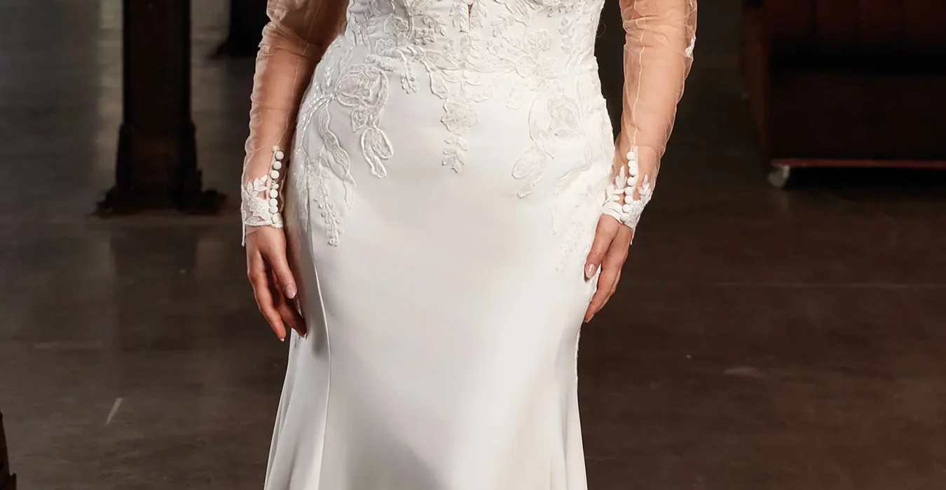 Eddy K fit-n-flare wedding dress with removable long sleeves, lace on bodice and end of train on bridal model standing with brown hair down.