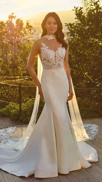Eddy K satin mermaid wedding dress with an illusion neckline and a sleeveless body on a bridal model standing in a garden with a sunset as a background.