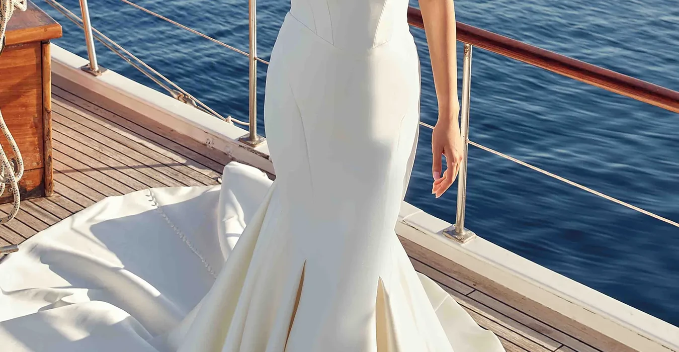 Eddy K strapless mermaid mikado wedding dress with sweetheart neckline photographed on bride standing on a boat on the ocean.
