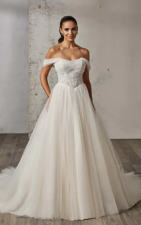 Eddy K tulle ballgown wedding dress with basque waist and off shoulder swag straps on bridal model standing in studio with hair pulled in a ponytail.