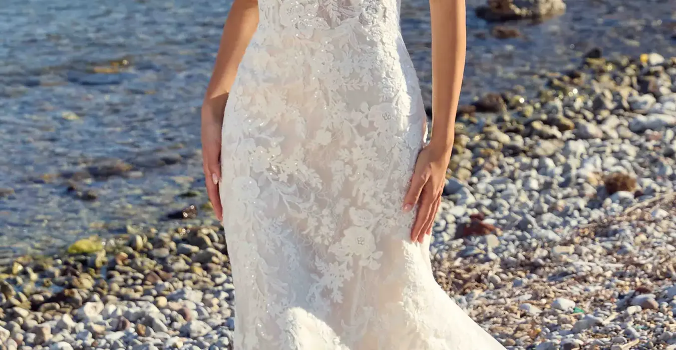 Eddy K lace fit-n-flare wedding dress with a straight neckline and a sleeveless body on a bridal model standing on a rocky beach.