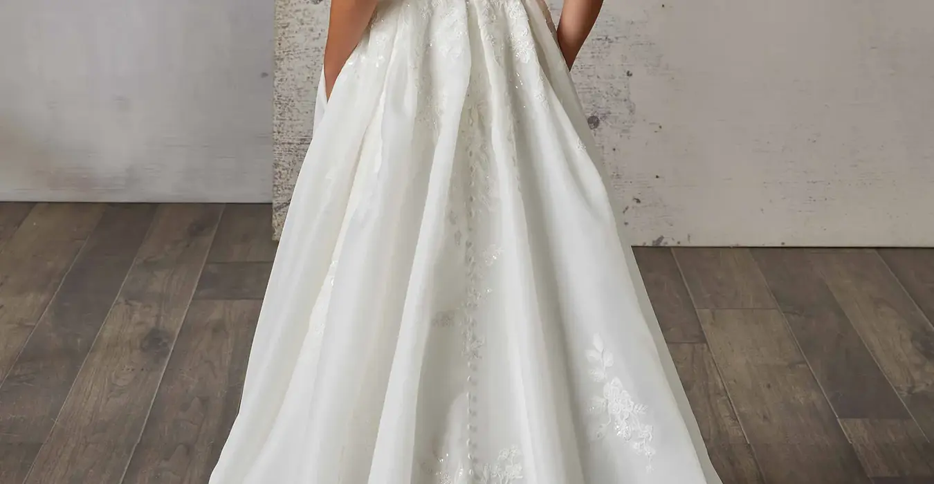 Back of Eddy K sleeveless ballgown with skin illusion lace back and organza skirt on bridal model standing in studio.
