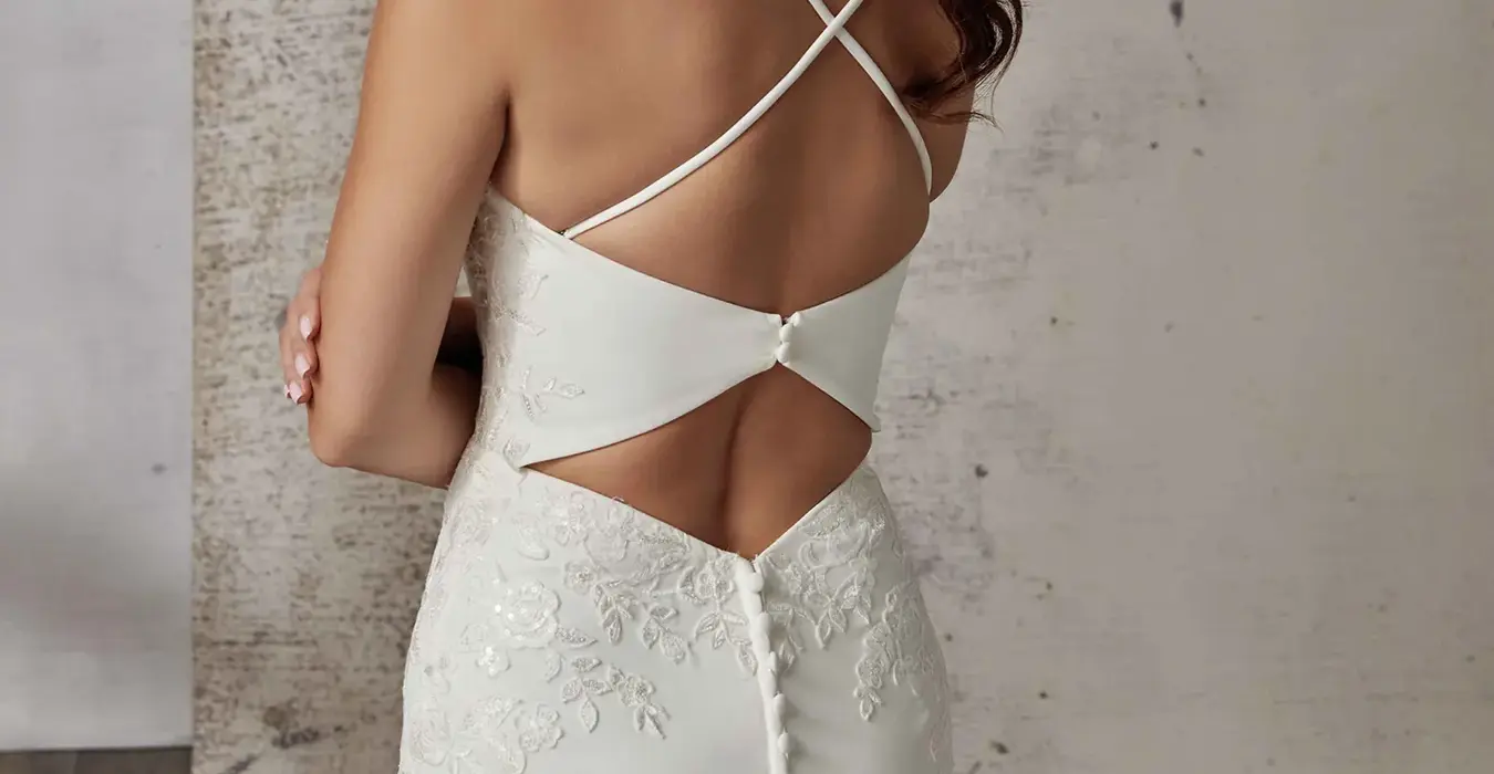 Back close up of Eddy K crepe wedding dress with keyhole back, criss crossed spaghetti straps and buttons down the trains on model standing in studio.