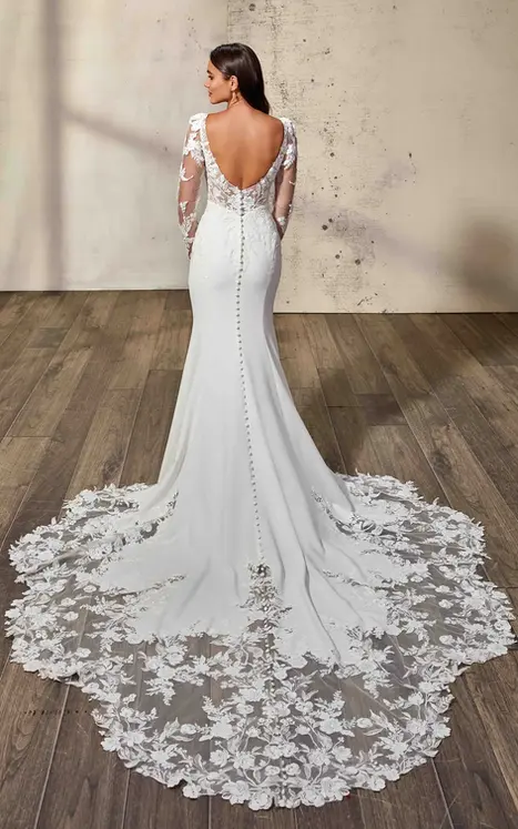 Back of Eddy K fit-n-flare crepe and lace wedding dress with illusion long sleeve and big lace train on bridal model standing in a studio.