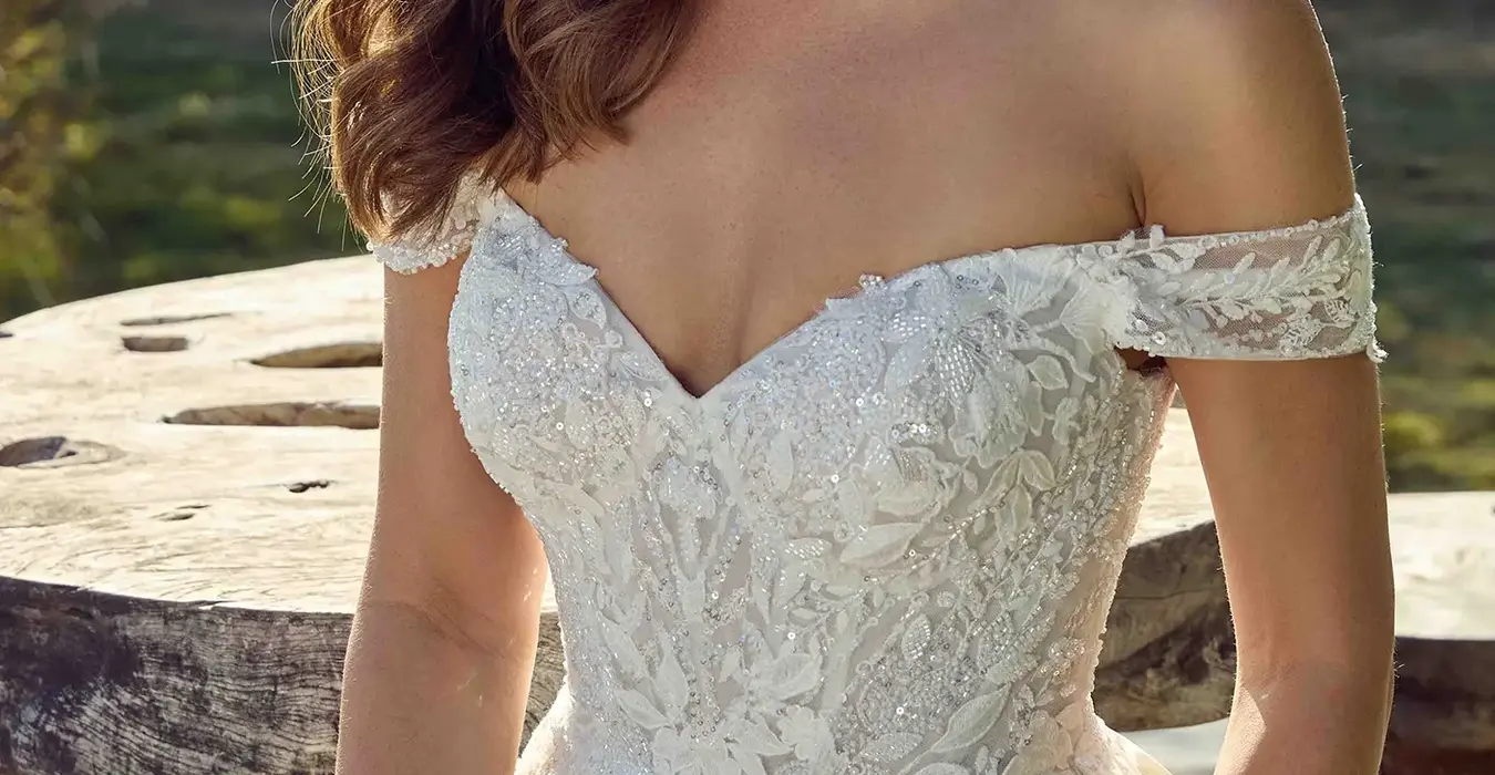 Close up of Eddy K mikado ballgown wedding dress with a sweetheart neckline and an off-shoulder body on a bridal model standing in a park in a sunny day with threes and green grass in the background.