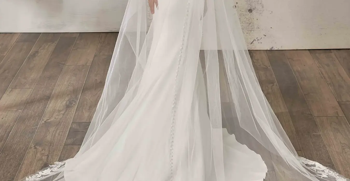 Back of Eddy K crêpe fit-n-flare wedding dress with a sweetheart neckline and an off-shoulder body on a bridal model standing in a bridal studio wearing along veil.