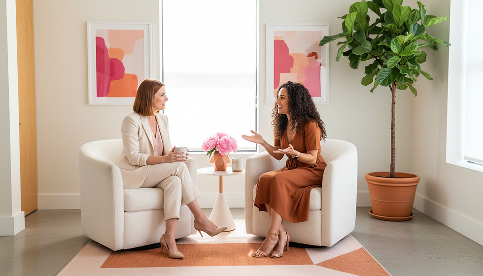 Two women sit in white chairs, talking and smiling, with pink flowers on a table. Abstract art and a potted plant decorate the room.