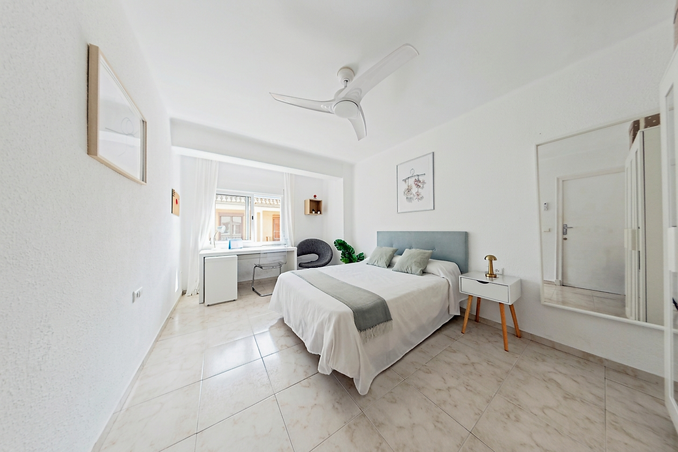 Interior design photography of a bright, modern bedroom with a minimalist aesthetic and a central double bed. The room features crisp white walls, a ceiling fan, and a workspace area, creating a calm and airy atmosphere.