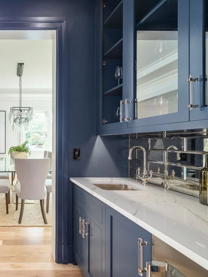 Symmetrical navy butler’s pantry with mirrored backsplash, white counters, and centered view into dining room.