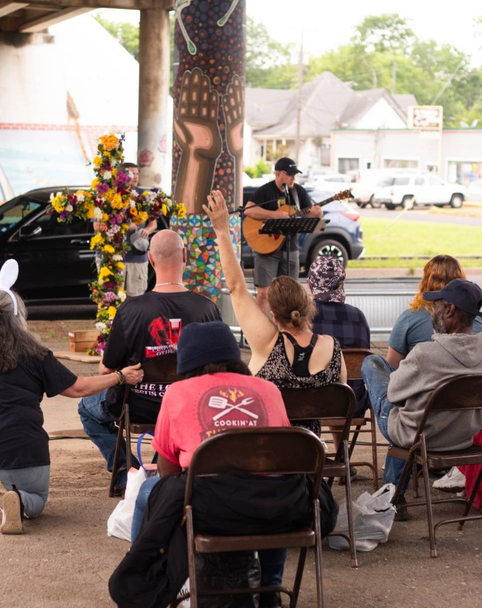 Community members seated under the Gentry Parkway bridge during a Hunger for Love Saturday Service. A musician plays guitar near a large cross decorated with flowers, while a woman in the crowd raises her hand in worship.