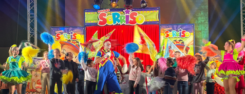Palco com luzes, cenário do espetáculo do Palhaço Sorriso mais as bailarinas e crianças com bandeiras a cantar. Mega espetáculo concerto do Palhaço Sorriso em Evento Infantil.