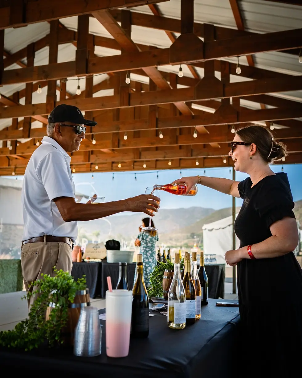 A woman pours wine for a man under a wooden pavilion. Bottles and greenery decorate the table. Mountains and a tent are visible in the background.