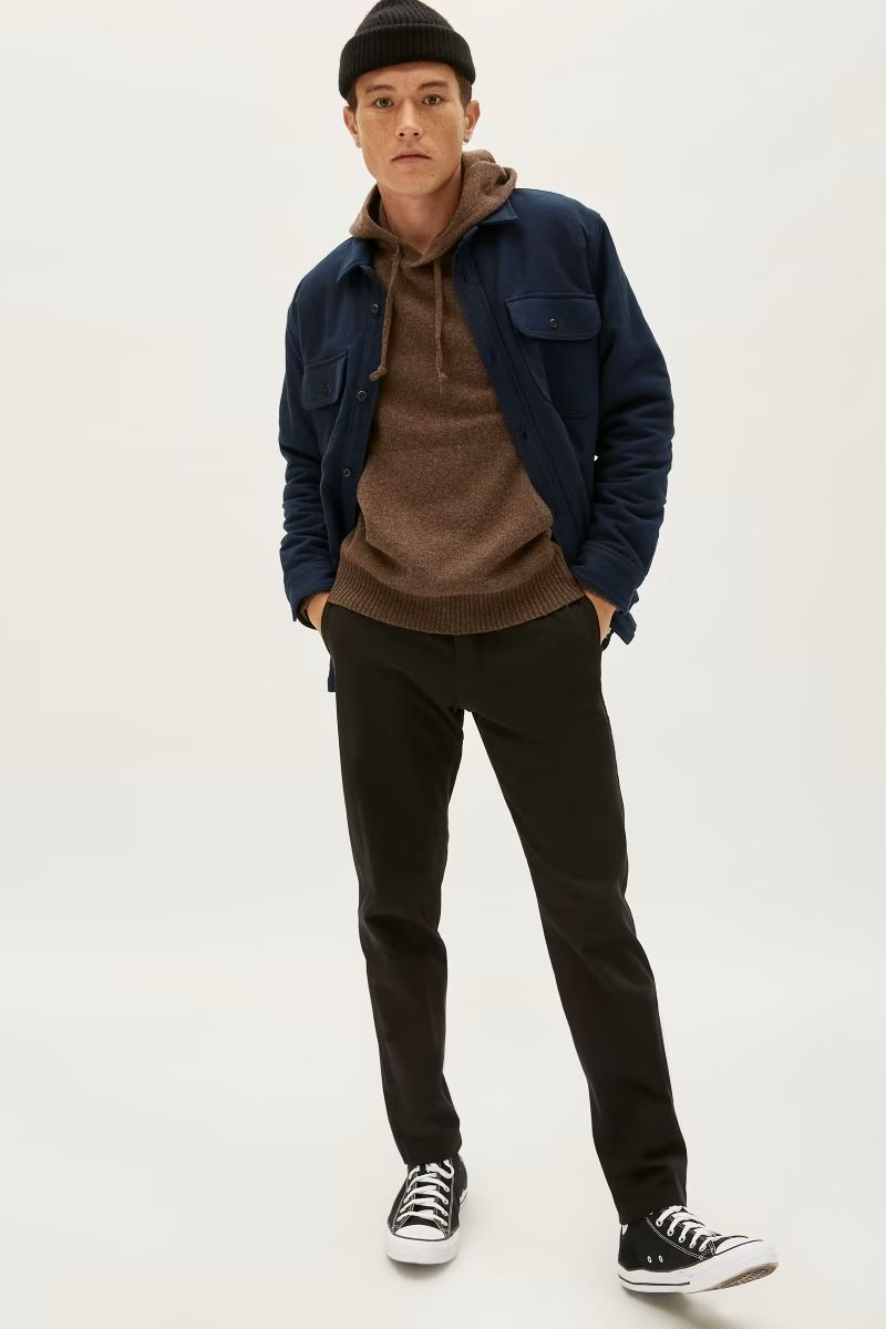 A person in a brown hoodie, blue jacket, black pants, and sneakers stands against a plain background, wearing a black beanie.