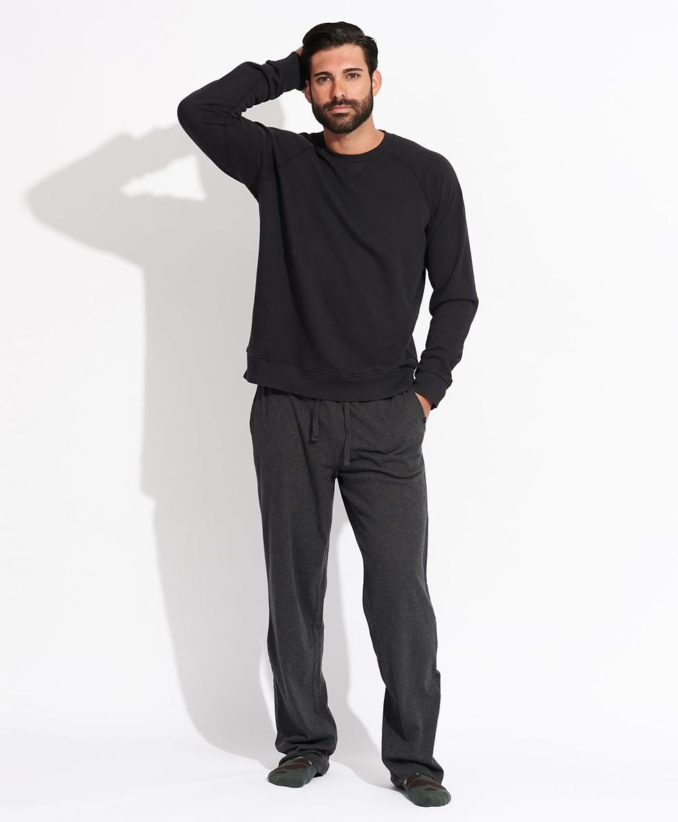 Man in a black sweatshirt and dark pants stands with hand on head against a white background, casting a shadow. Neutral expression.