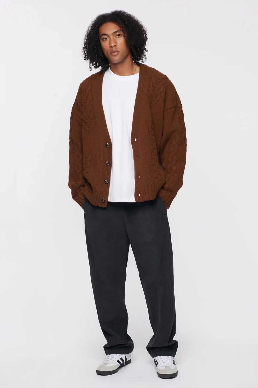 Person in a brown cardigan and white shirt stands against a plain white background, wearing black pants and white sneakers, looking relaxed.