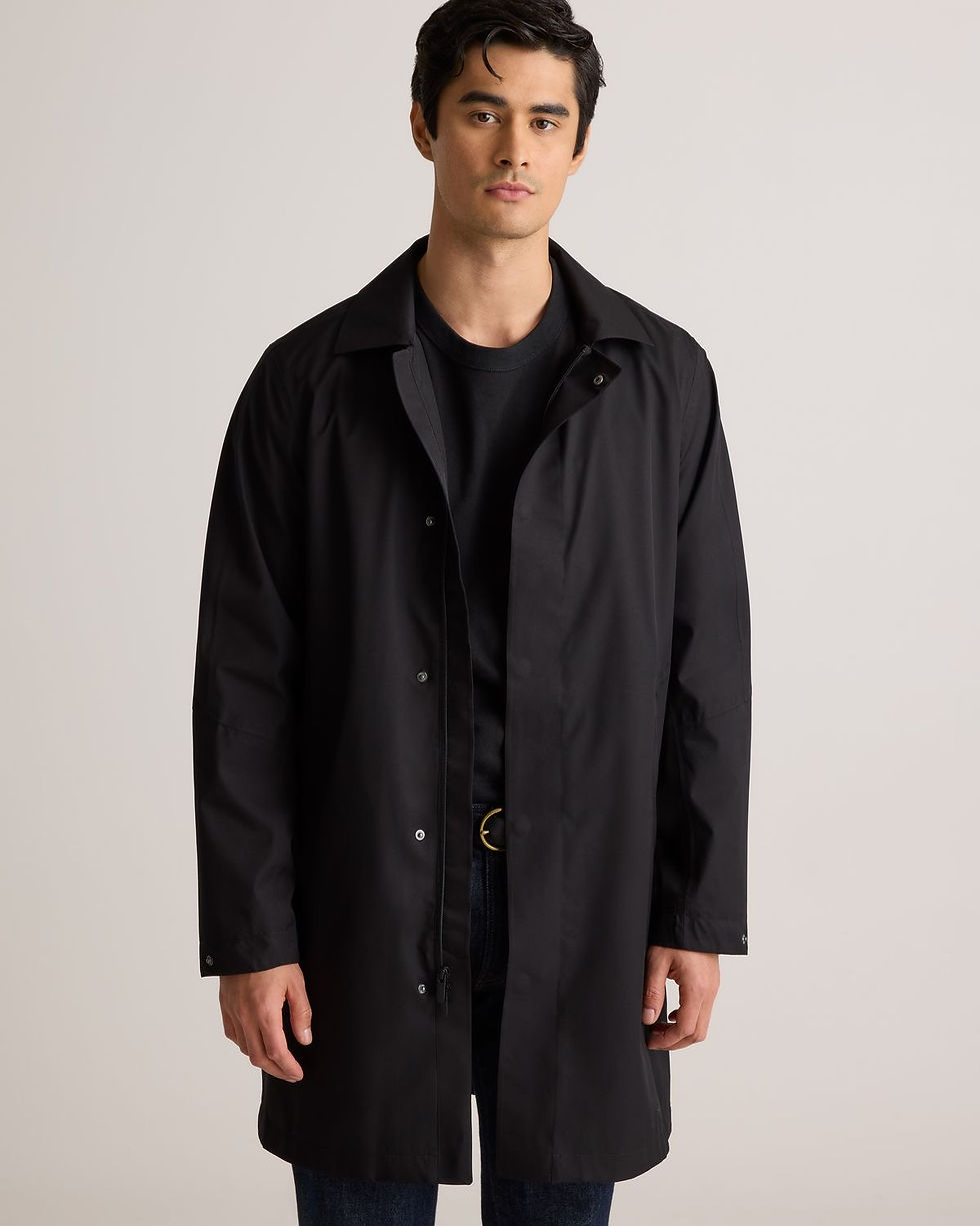 Man in a black coat stands against a neutral background, looking calm. He's wearing a black shirt and jeans, with no visible text.