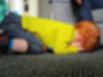 Young child with red hair in a yellow shirt kneeling on a gray carpet, face down. Cozy indoor setting with an adult's hand nearby.