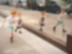 Two young boys joyfully run alongside a glass wall, showing their reflections. They hold leaves, with parked cars in the background.