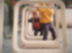 Two kids play joyfully through geometric frames in a park. One wears orange and gray, the other in yellow. Bikes and plants are visible.