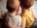 Two toddlers sit closely together, one in a gray shirt and the other in yellow, on a patterned rug. They show a sense of companionship.