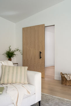 A cozy living room corner with a white sofa adorned with striped pillows. The room features a wooden door with a detailed pattern, light wood floors, and a small plant, creating a warm and inviting atmosphere.