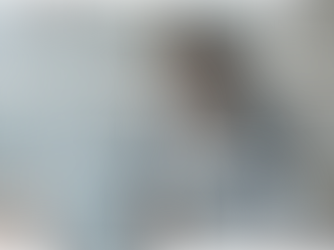 Mulher sentada em um escritório individual, digitando em um notebook sobre a mesa. Ela mantém postura reta, com o olhar levemente cabisbaixo, expressão de desânimo contido. Há papéis espalhados na mesa e uma janela ao fundo, em um ambiente sereno e silencioso.