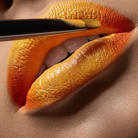 Extrem makrobild inom skönhetsfoto där en professionell sminkborste applicerar ett läppstift med gul och orange ombre-effekt på en kvinnas fylliga läppar, visar vita tänder. Kommersiell high-fashion kosmetikareklam och sminkkampanj av Nello Johnson Photography Katrineholm Sverige.