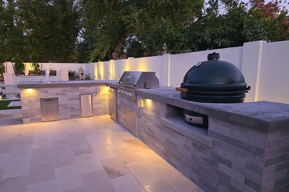 Outdoor kitchen with a grill and a green ceramic cooker on a stone counter. Soft lighting, white fence, and trees in the background.
