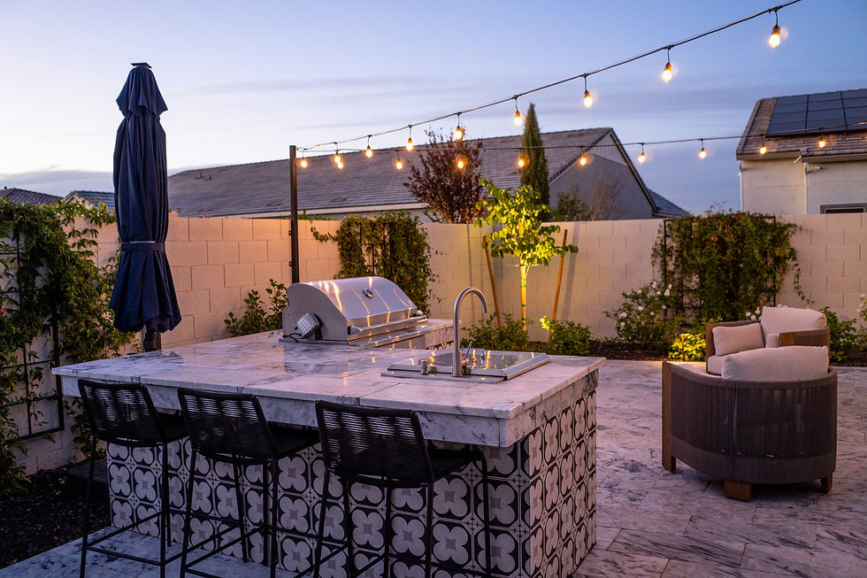 Outdoor kitchen with marble counter, grill, and sink. String lights illuminate the tiled pattern. Patio chairs and plants create a cozy vibe.